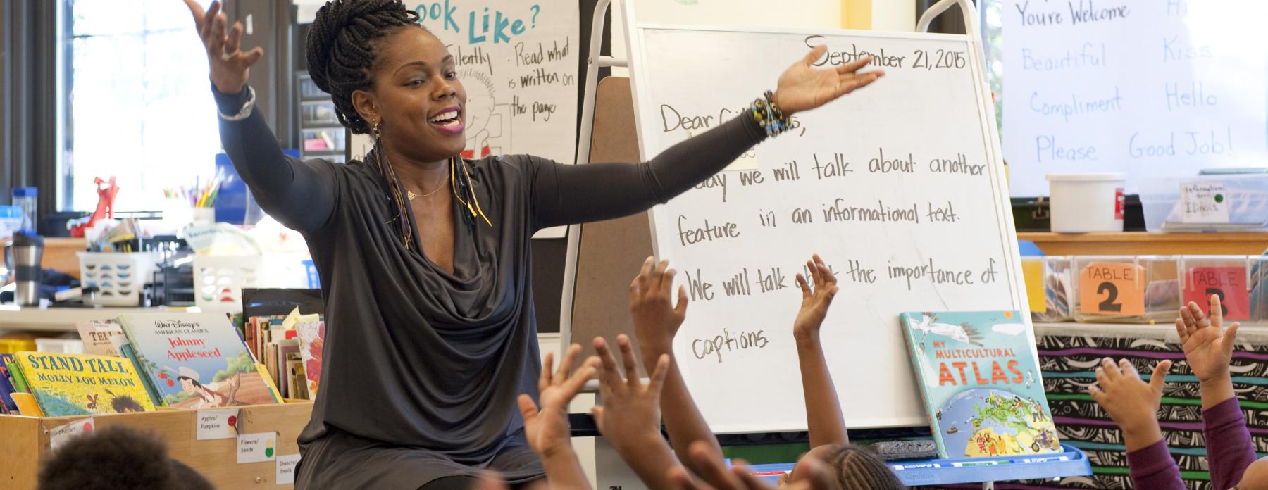 An elementary school teacher extends her arms into the air as she leads her students in song